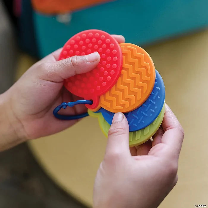 Hands hold four round silicone mats in red, orange, blue, and lime green with varying raised textures on a blue clip.