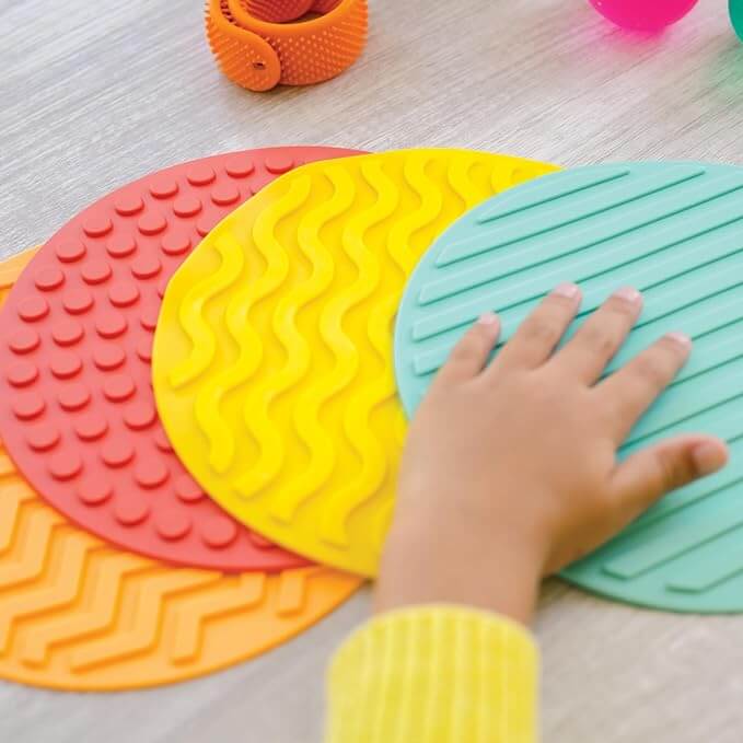 Four colorful circular silicone mats with various raised textures; a child's hand touches the ridged teal mat.