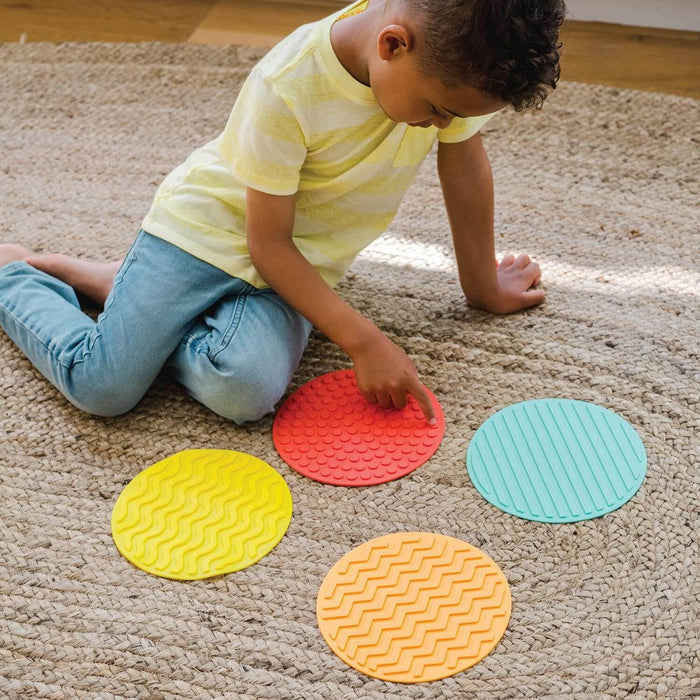 A boy touches four colorful, textured circular mats: yellow waves, red dots, orange zigzags, and blue lines.