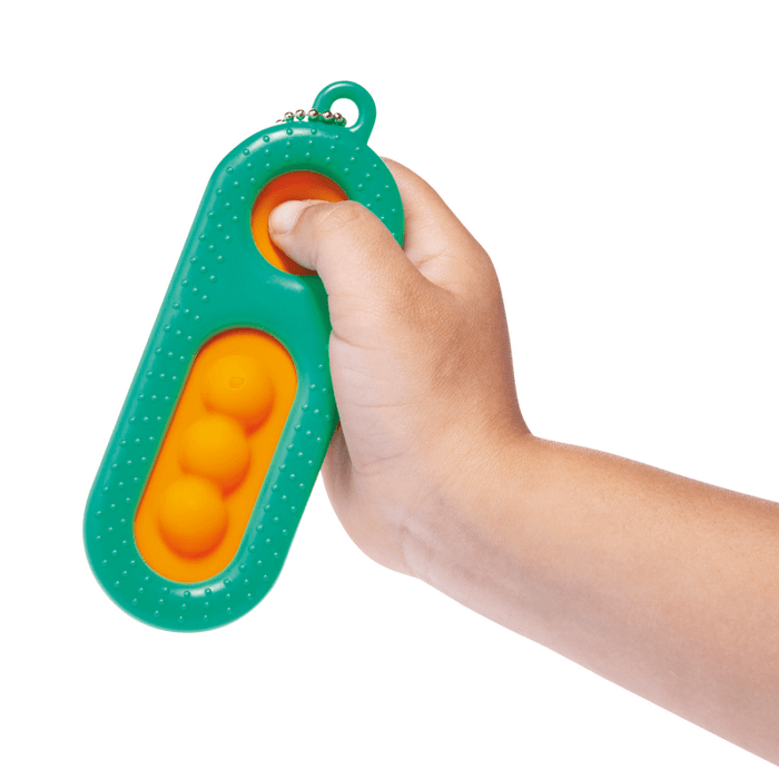 Hand holds a green textured pill-shaped fidget toy with orange popping bubbles. A thumb presses down on one bubble.