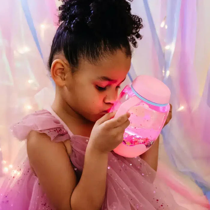 Young girl holding a glowing, round pink sensory jar with glitter and a pink lid, wearing a pink ruffled dress.