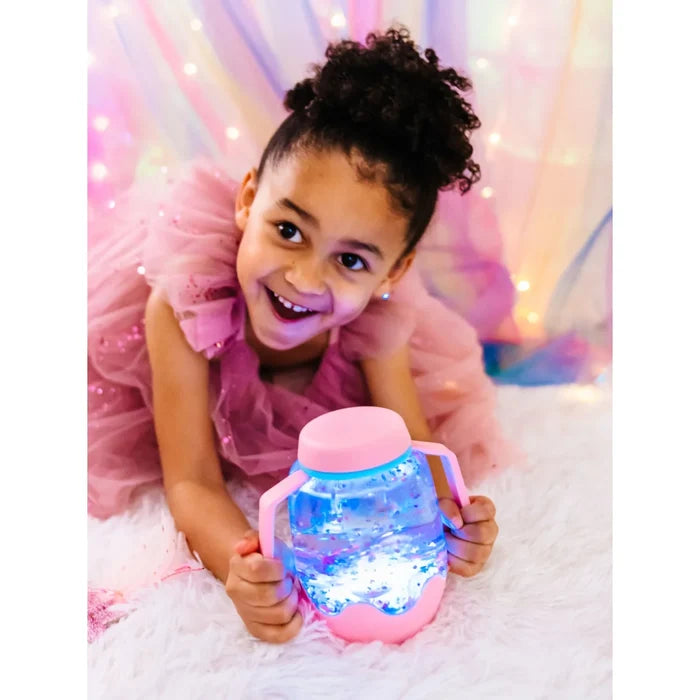 Smiling girl holds a pink sensory jar with glowing blue water and glitter, featuring pink handles and lid.