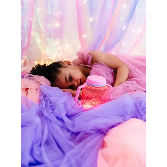Child lying in pink and purple tulle looks at a pink glowing sensory jar filled with swirling glitter.