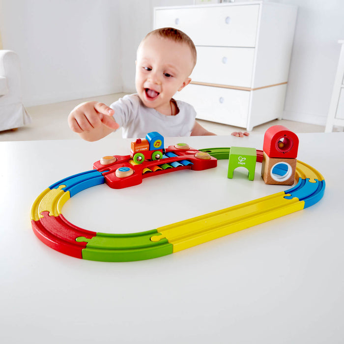 Toddler playing with a colorful wooden train set featuring a musical bridge and sensory blocks on an oval track.