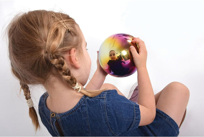Child holding a large, iridescent reflective sphere in yellow and pink, viewing their reflection in its shiny surface.