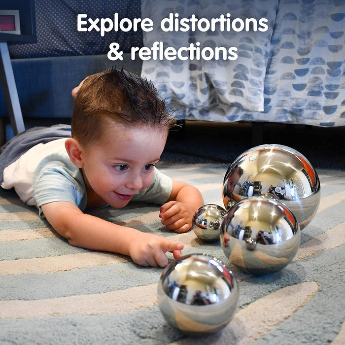 A young boy plays with four reflective, mirror-finish silver spheres of various sizes on a striped rug.