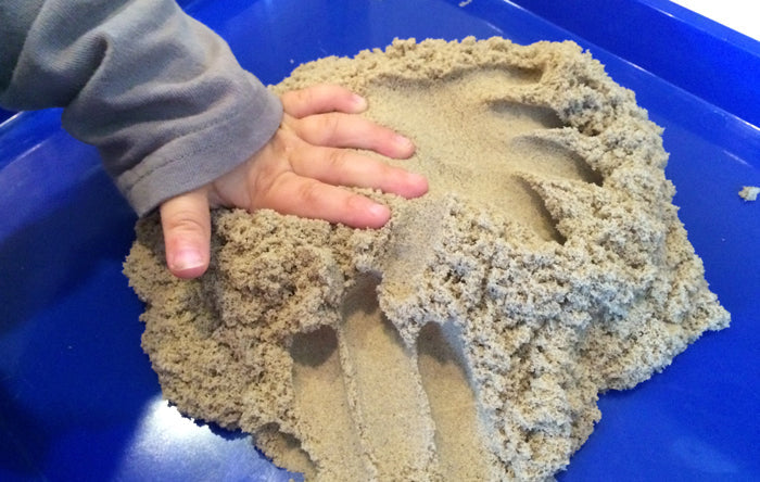 Child's hand pressing into tan moldable sand on a blue tray, creating smooth finger tracks and textured mounds.