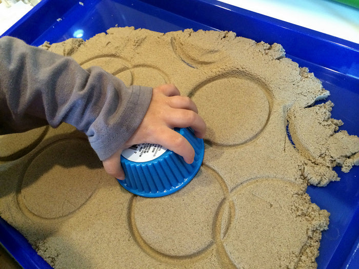 Child's hand pressing a blue cup into tan moldable sand to create circular imprints in a blue tray.
