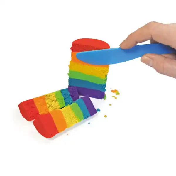 Hand using a blue knife to slice a rainbow-layered cylinder of colorful sensory sand into segments.