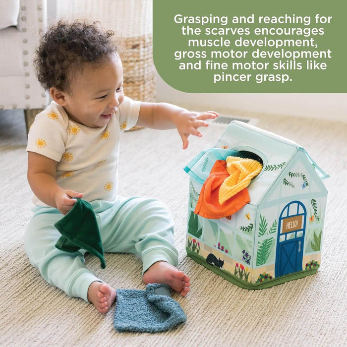 Baby reaching for colorful fabric scarves inside a soft house-shaped toy box on a rug.