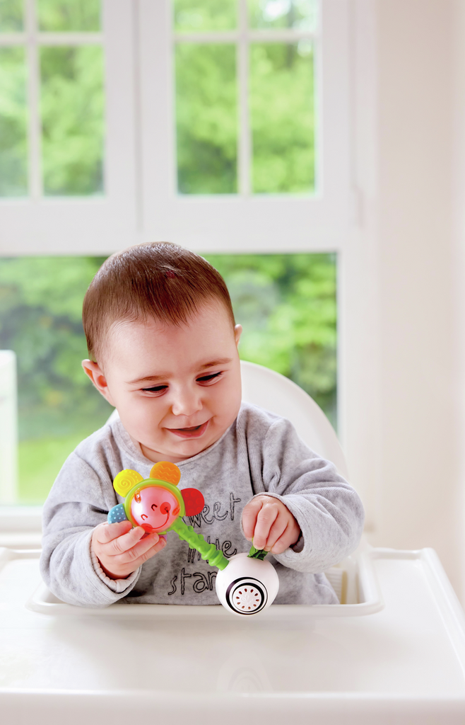 Baby in a high chair holds a flower rattle with colorful petals, a green textured stem, and a white round base.