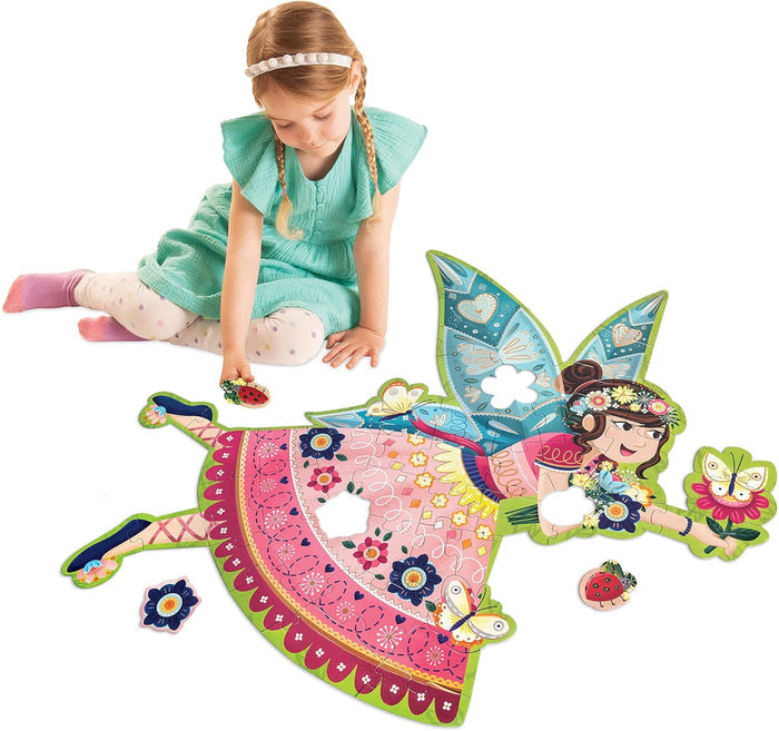 Child assembling a large shaped floor puzzle of a fairy with shimmering blue wings and a patterned pink dress.