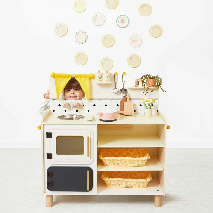 Cream wooden play kitchen with a microwave, wicker baskets, and accessories. A child peeks through a yellow window.