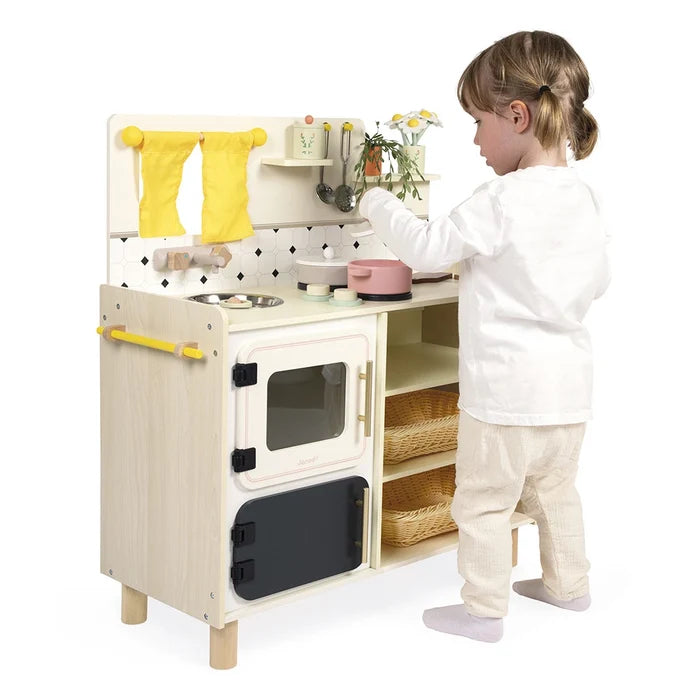 Child playing with a light wood toy kitchen featuring yellow curtains, white oven, and wicker storage baskets.