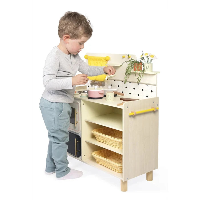 Young boy playing with a light wood toy kitchen featuring yellow accents, wicker baskets, and a tiled backsplash.