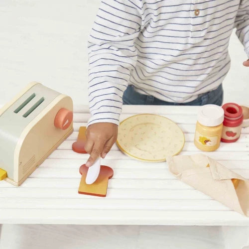 Child playing with a sage green and cream wooden toy toaster set including toast, jars, a plate, and a white toy knife.