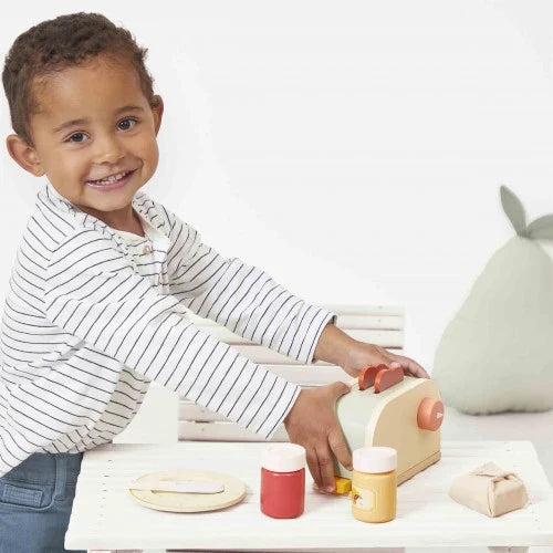Child playing with a mint and wood toy toaster, colorful jars, and a wooden plate set on a white table.