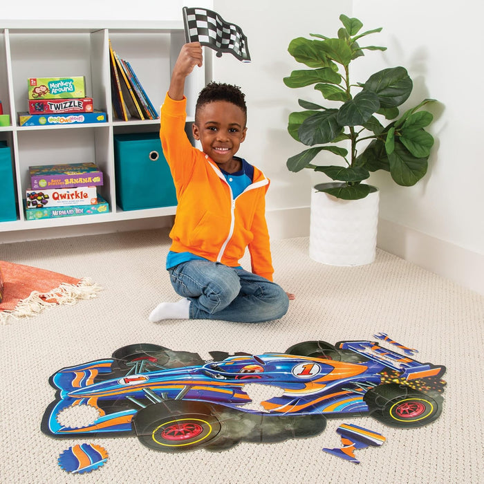 A boy holding a checkered flag sits behind a large, blue and orange racecar-shaped floor puzzle on a cream carpet.