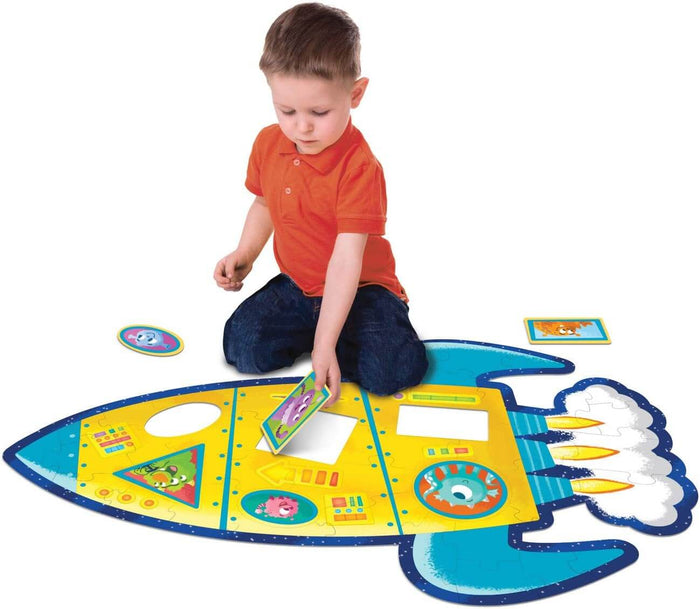 Boy assembling a large yellow and blue rocket-shaped floor puzzle with colorful cartoon alien illustrations.