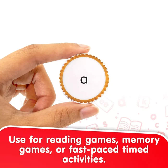 A hand holds a round snack-shaped token with a scalloped orange border and the letter a on its white center.