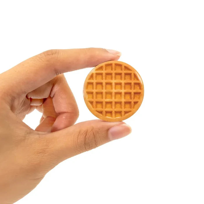 Hand holding a miniature round tan waffle toy with a grid pattern against a white background.