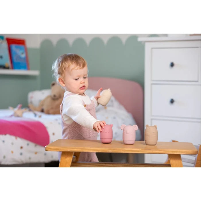 Toddler plays with four pastel pink and beige animal-shaped silicone toys on a wooden table in a nursery.