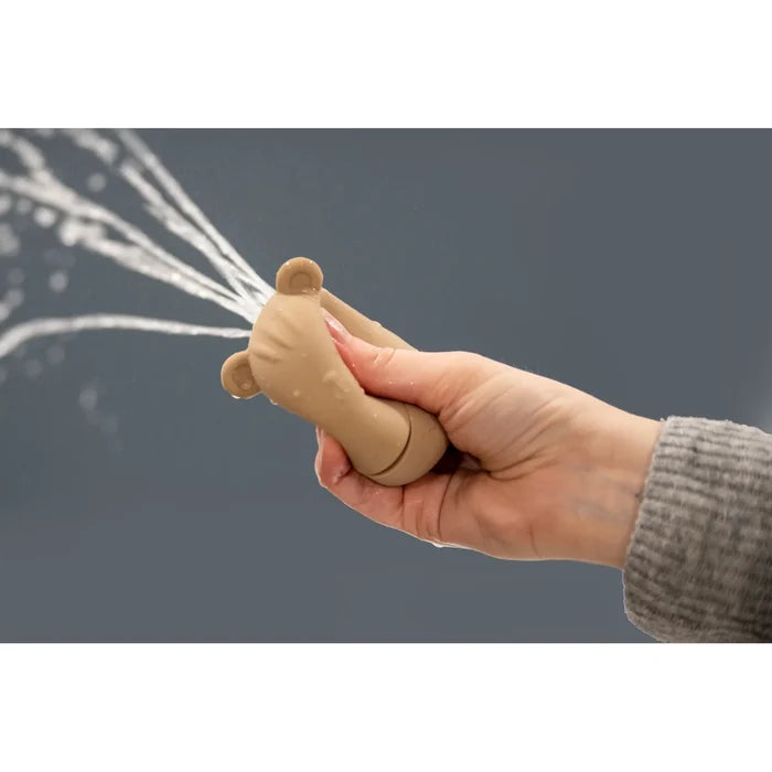 Hand squeezing a tan silicone bear-shaped bath toy, spraying multiple jets of water against a gray background.