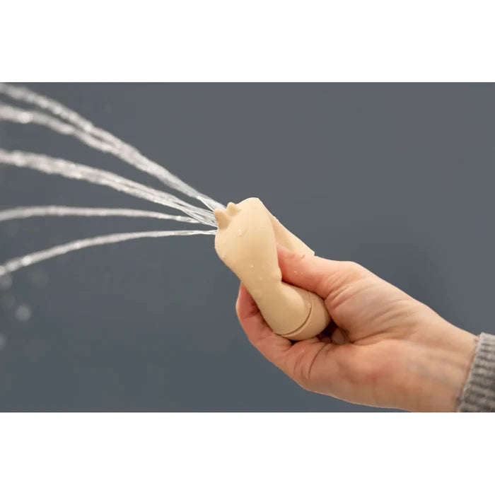 A hand squeezes a tan silicone animal toy, spraying several streams of water against a solid grey background.