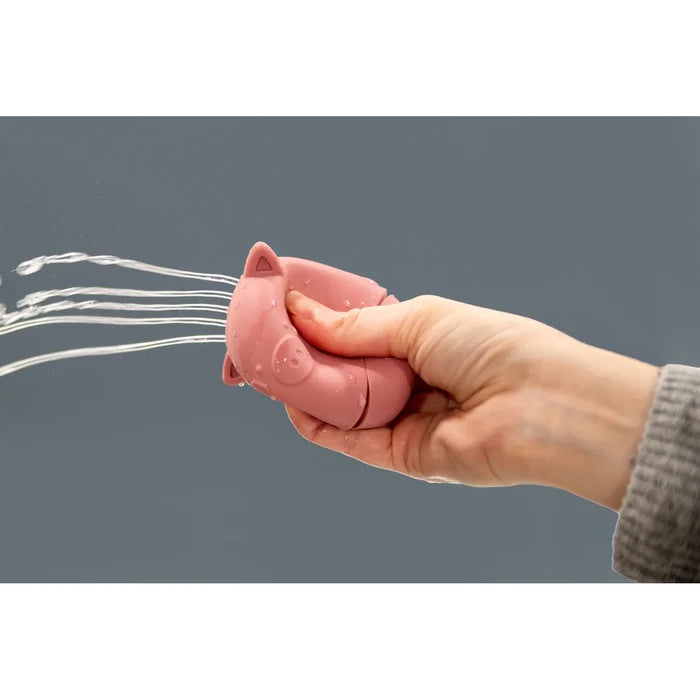 A hand squeezes a pink pig-shaped silicone toy, spraying multiple streams of water from its snout.
