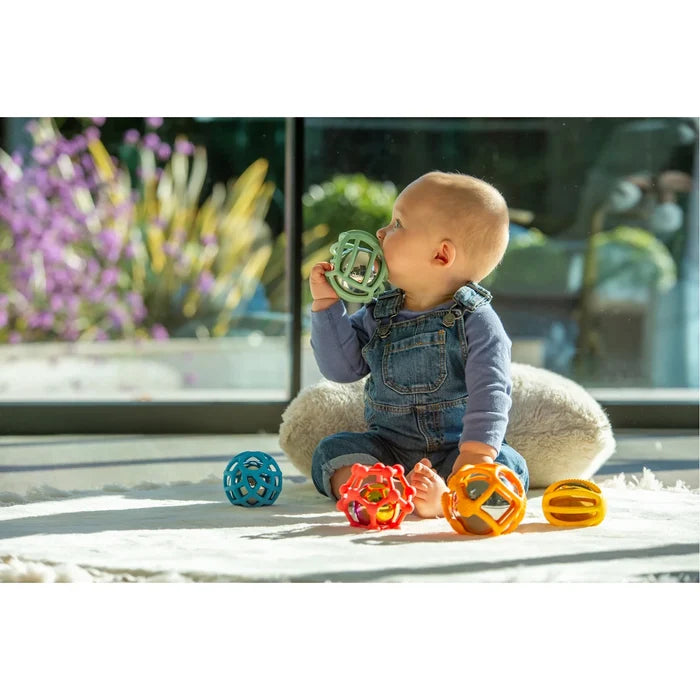 Baby sits on a rug with five colorful, flexible, geometric balls in blue, red, orange, yellow, and mint green.