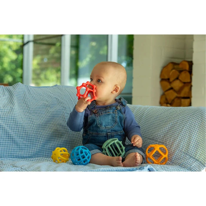 Baby on a blue checkered couch holding a red flexible ball to its mouth, with four other colorful balls nearby.