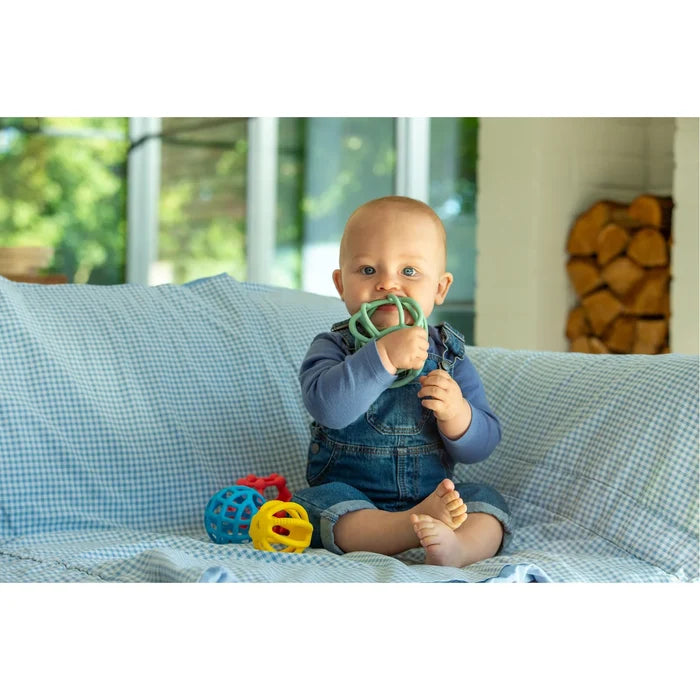 Baby on a blue checkered couch biting a green flexible ball with red, blue, and yellow cage-like balls nearby.