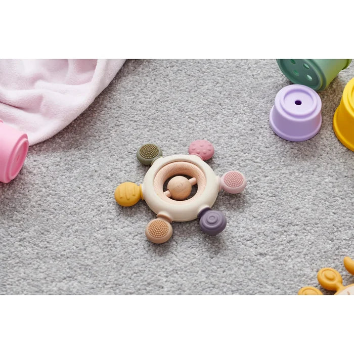 Round baby teether with earth-toned textured silicone nubs and a wooden rattle center, shown on a grey carpet.