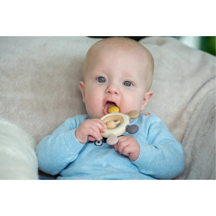 A baby chewing on a round wooden rattle teether featuring four textured silicone nubs in muted earth tones.