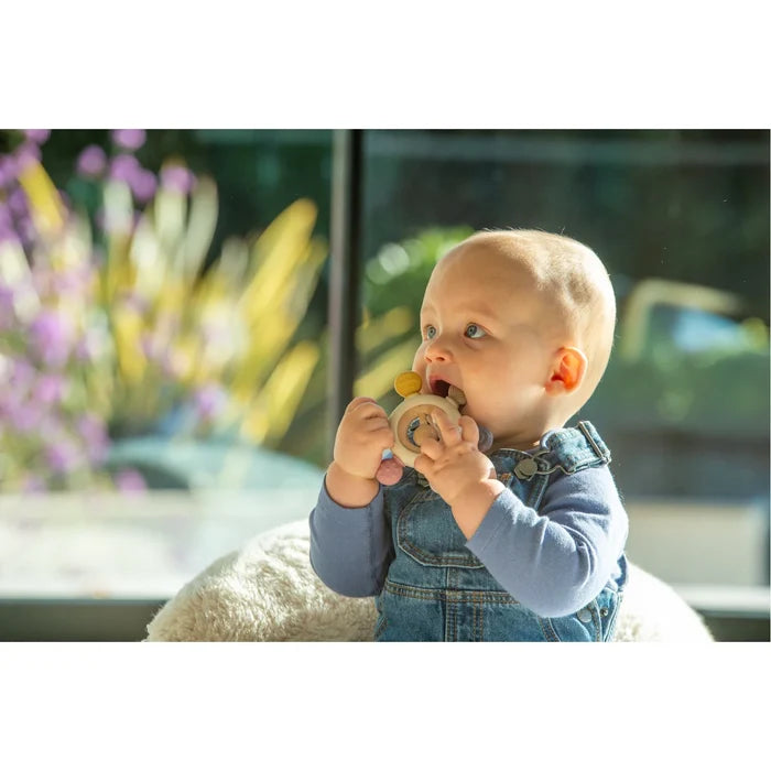 Baby holds a circular wooden and silicone teether with mustard and pink accents and a spinning center element.