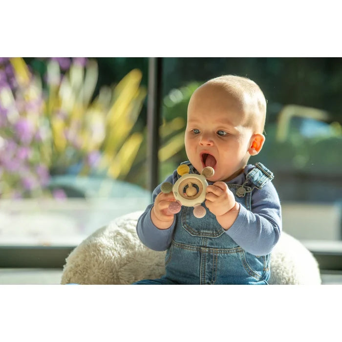 Baby holding a sun-shaped silicone teether with earth-toned textured nubs and a central wooden rattle.