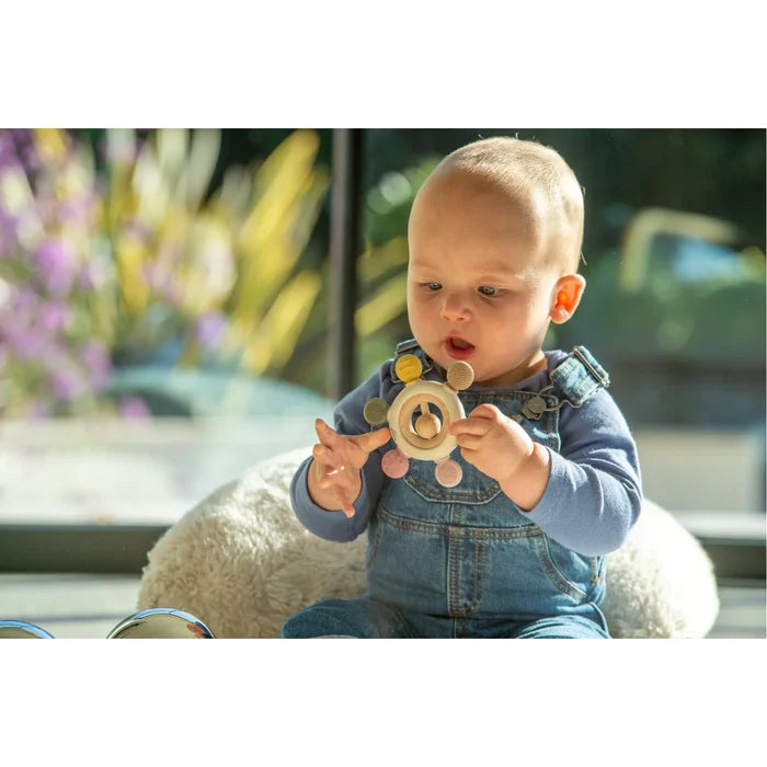 Baby in denim overalls holding a round wooden and silicone teether with colorful textured knobs and a center rattle.