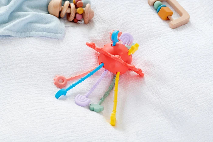 Red spherical baby toy with colorful textured silicone pull strings in pastel shades on a soft white blanket.