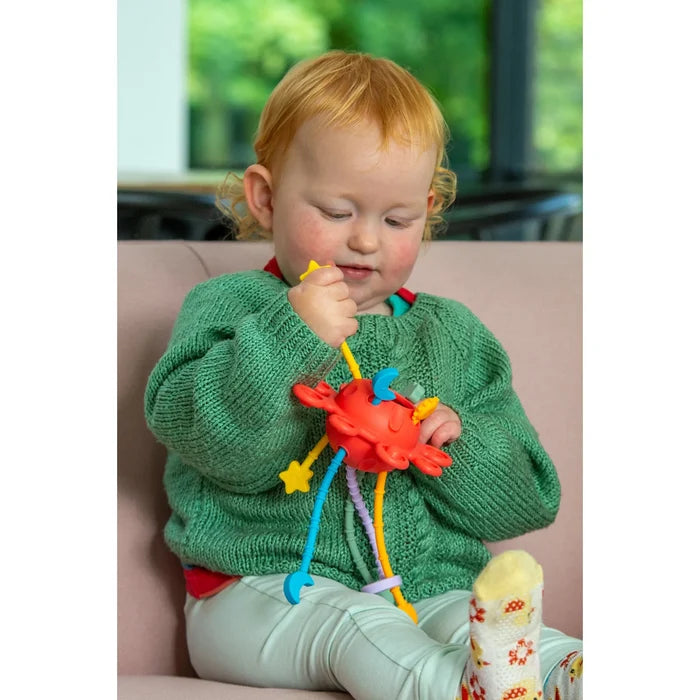 Toddler pulls on a red sensory toy with various colorful, textured silicone cords ending in star and moon shapes.
