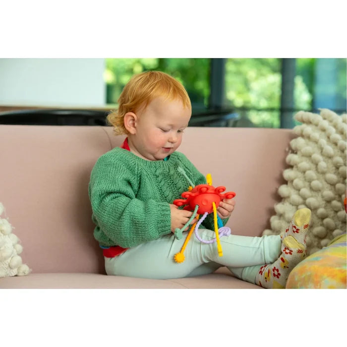 Toddler on a sofa playing with a red circular toy featuring colorful, textured sensory pull strings.