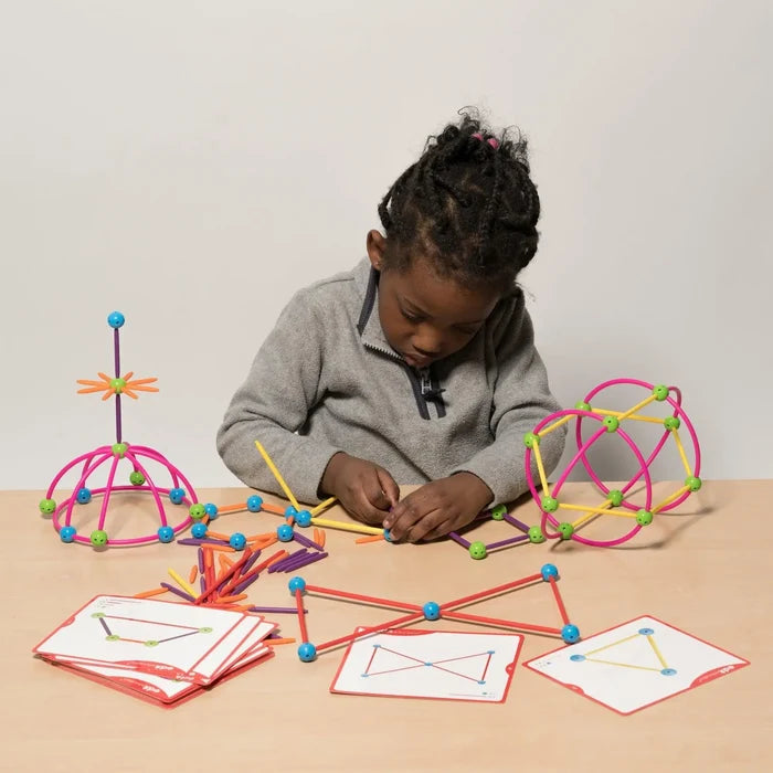 Child builds colorful 3D geometric structures using plastic rods, sphere connectors, and instruction cards on a table.