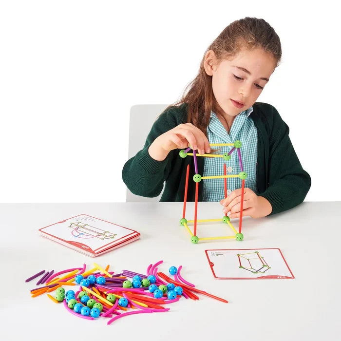 Girl building a 3D structure with colorful plastic rods and green connector spheres on a table with instruction cards.