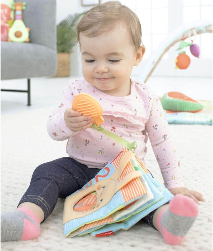 Baby sits on a rug playing with a colorful fabric activity book and its attached orange leaf-shaped teether.