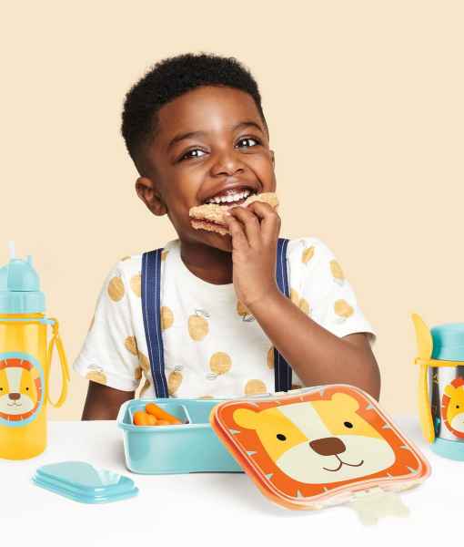 A child eats beside a blue lunch kit with an orange lion face lid and a matching yellow lion water bottle.