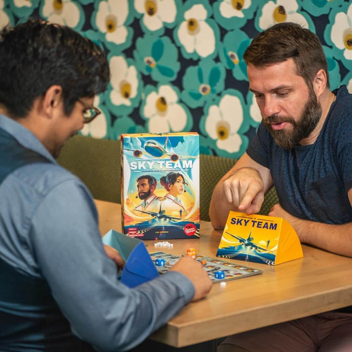Two men play Sky Team board game with an airplane-themed box, yellow triangular screen, and colorful dice on a table.