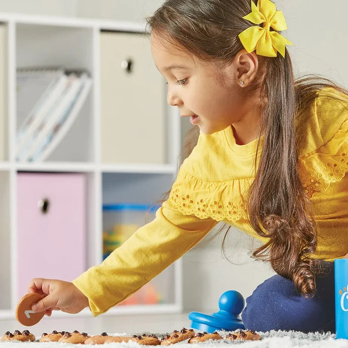 A girl in a yellow shirt playing with numbered toy chocolate chip cookies lined up on a white rug.