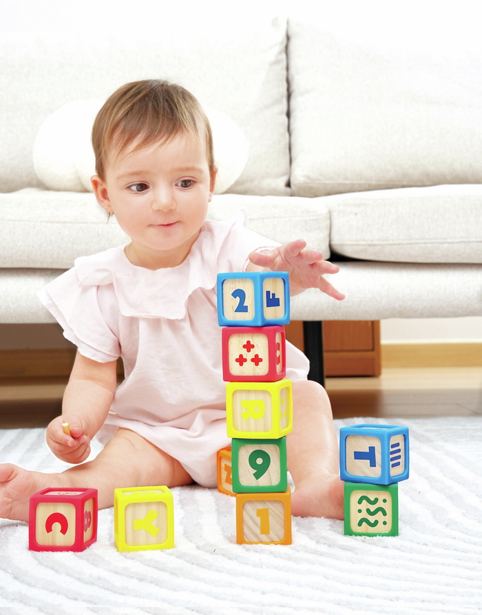 Baby stacking colorful wooden blocks with soft rubber edges and printed letters and numbers on a white rug.