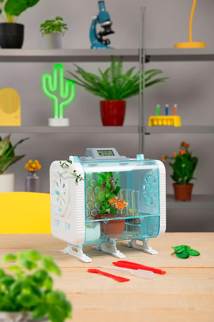 White and blue toy greenhouse with a digital display and plants inside, sitting on a wooden table with tools.