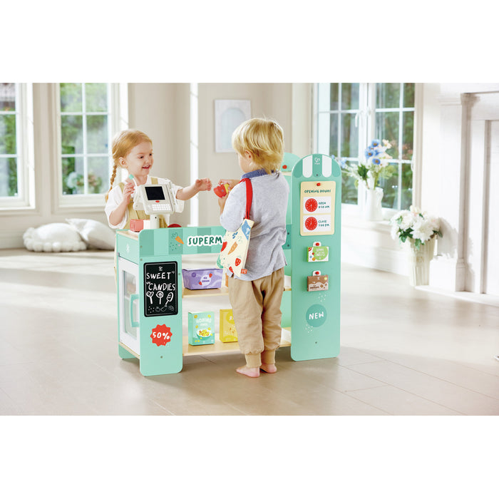 Two children play with a mint green wooden toy supermarket featuring a cash register, shelves, and colorful accessories.