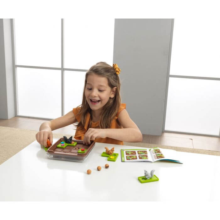A child plays with a squirrel logic puzzle featuring green tiles, brown acorns, and a brown game board on a white table.